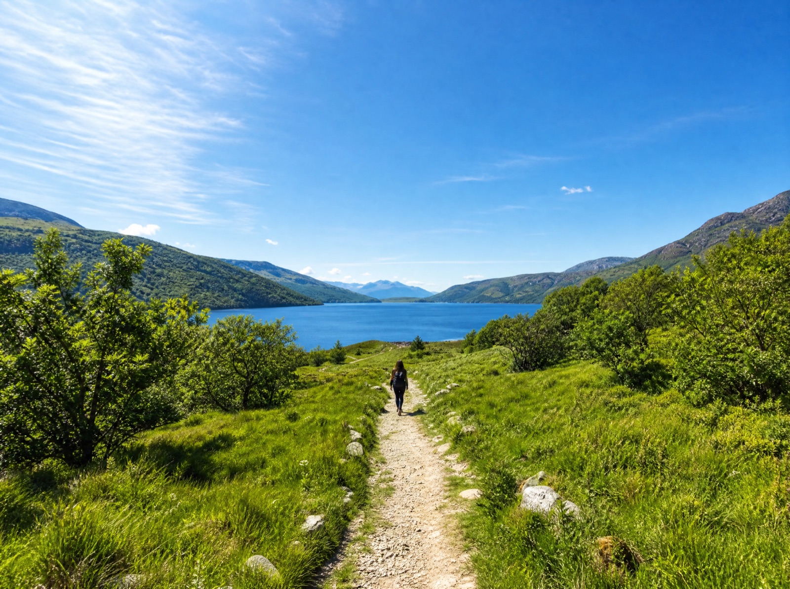 Eine kleine Person, von hinten und weit entfernt, geht auf einem hellen Weg durch grüne Natur zu einem See unter blauem Himmel.